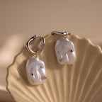 The Celestial Baroque Pearl Charm Hoops, featuring silver statement hoops with irregular pearl charms and small gems, are displayed on a shell dish for the perfect baroque look.
