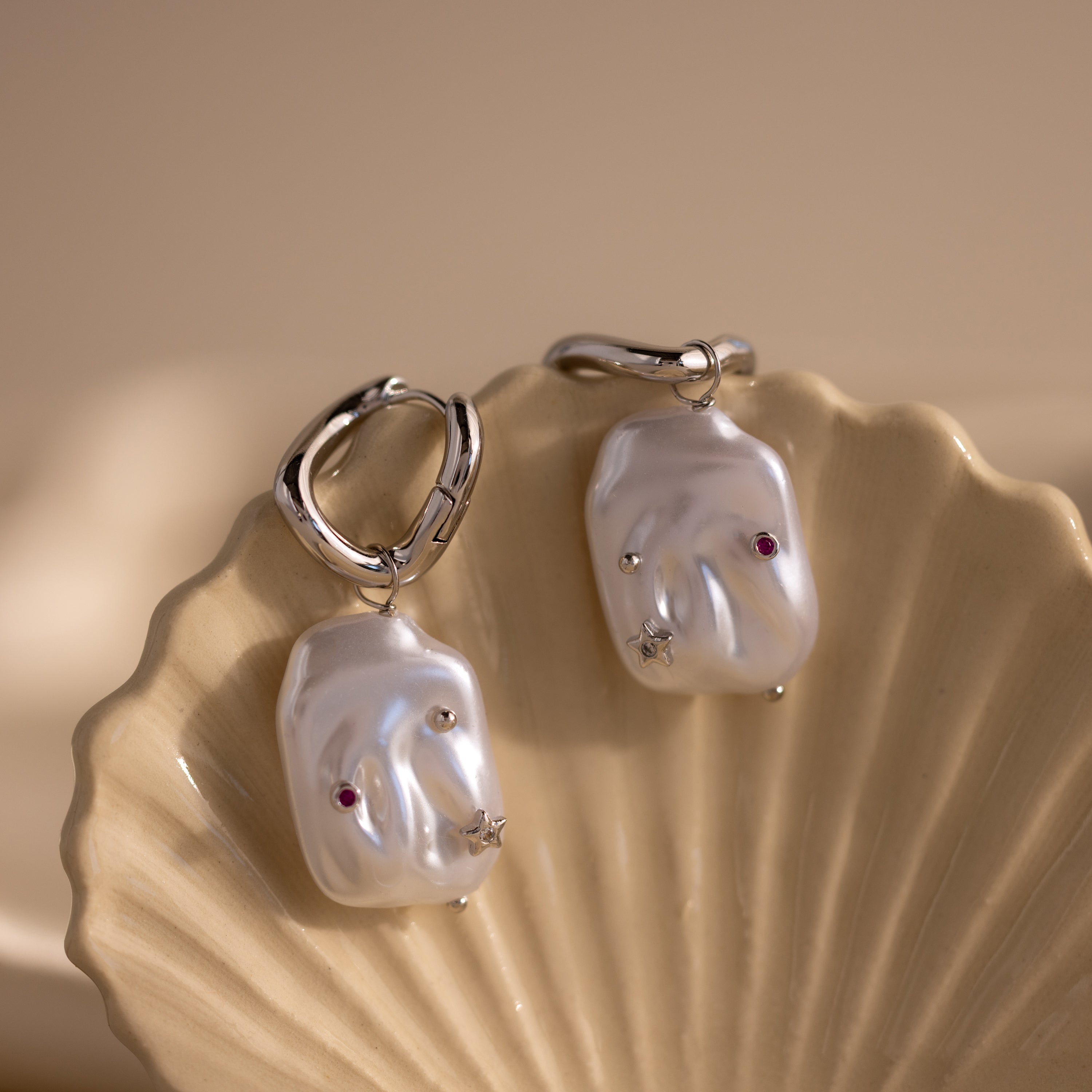 The Celestial Baroque Pearl Charm Hoops in White Gold, adorned with star and gemstone accents, are displayed on a shell-shaped dish.