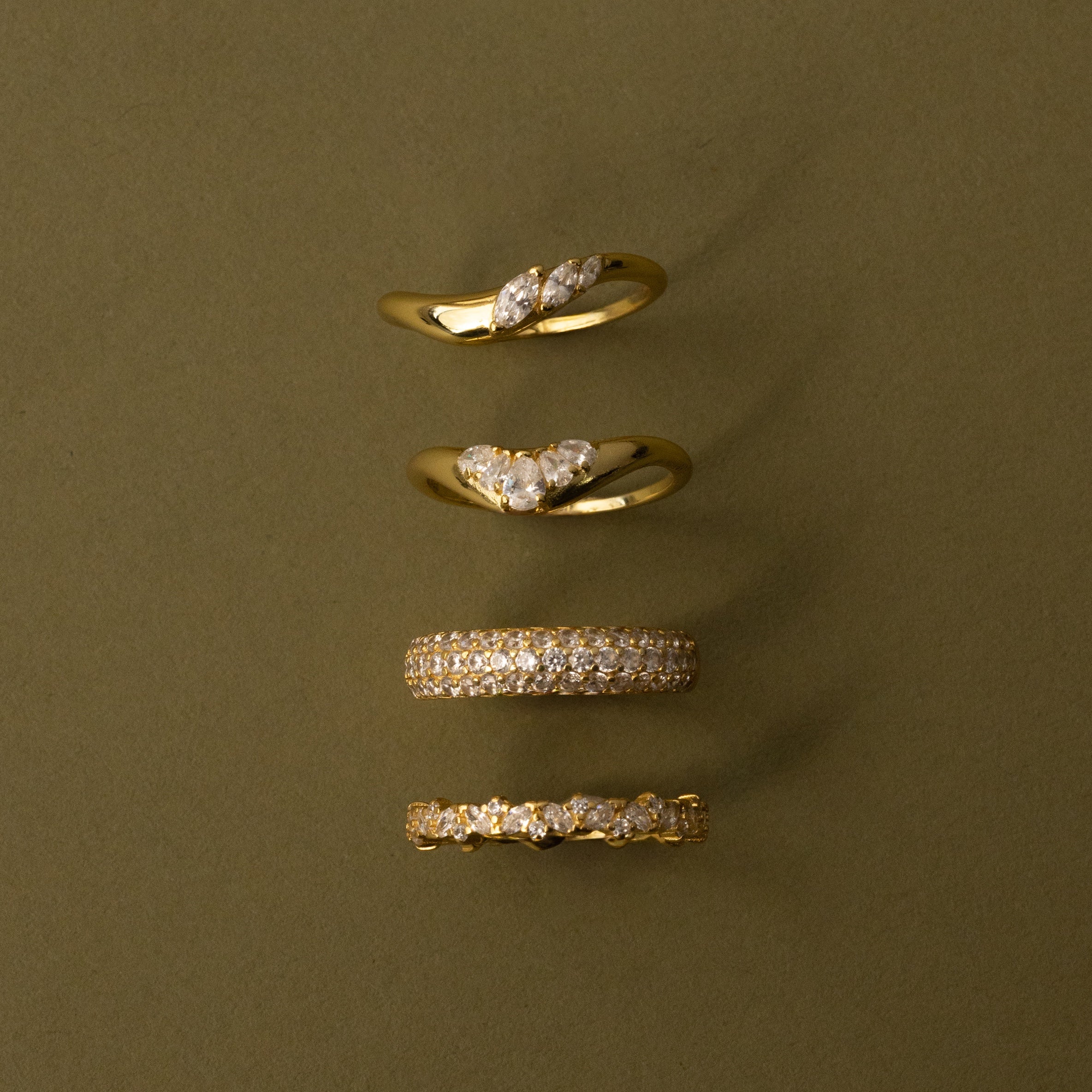 The Mixed Metal Diamond Curve Ring Set, featuring four stacked rings with diamond accents, is displayed vertically against an olive green background.