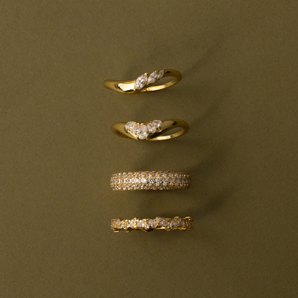 The Sunrise Diamond Curve Ring and three other gold rings with diamond accents are displayed vertically on an olive-green background for a modern, minimalist look.
