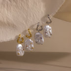 Two pairs of Celestial Baroque Pearl Charm Hoops—one gold and one silver, each with irregular baroque pearl drops—are elegantly displayed on a beige background.