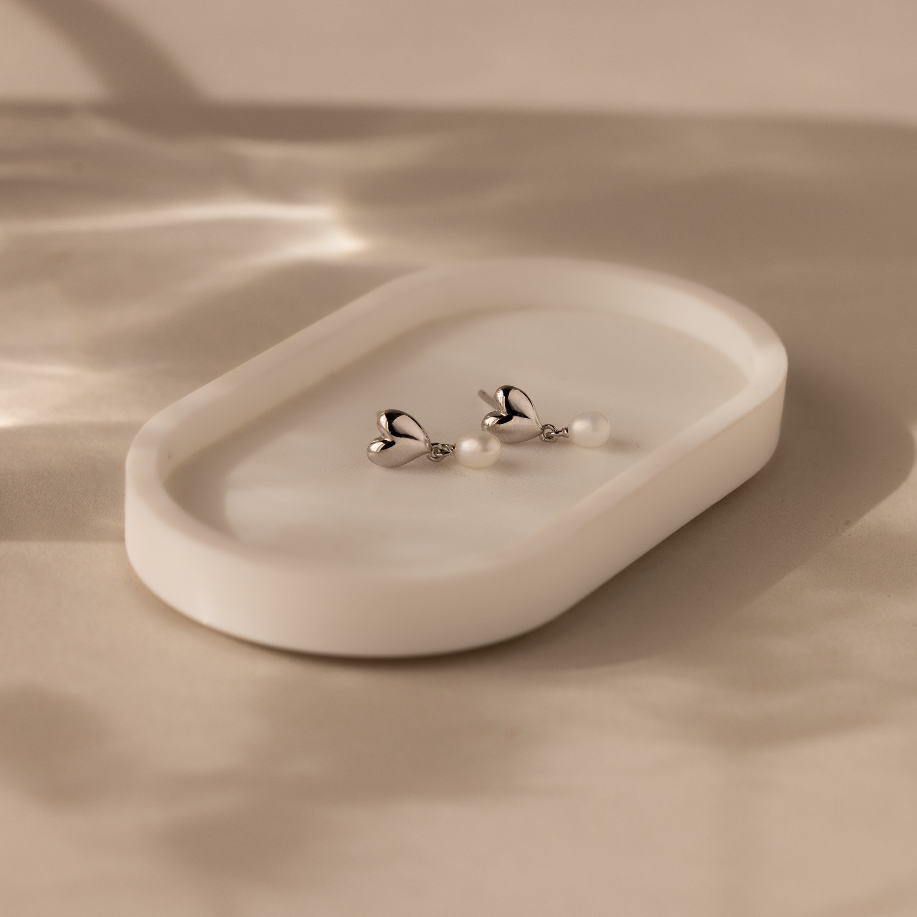 The Heart Pearl Drop Earrings in Silver are displayed on a white oval dish, with soft shadows and light highlighting their elegant silver and pearl accents.