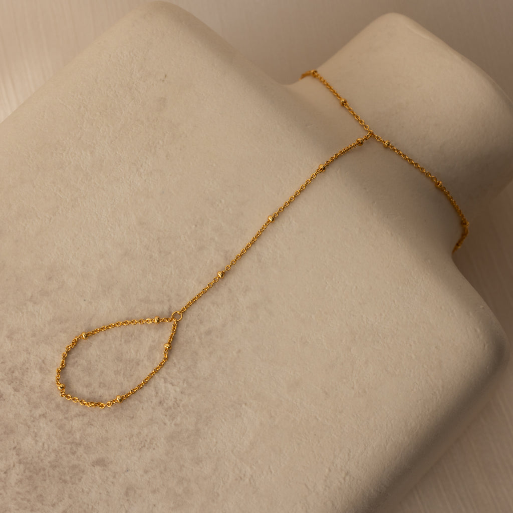The Beaded Hand Chain Bracelet in 18K Gold, displayed on a beige textured surface, is a stunning statement piece that pairs beautifully with a delicate chain ring.
