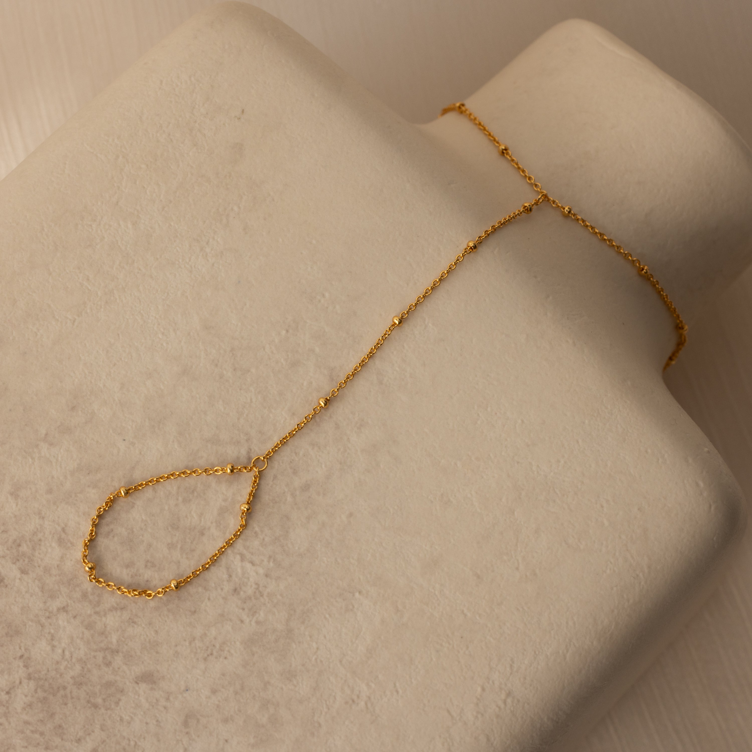 The Beaded Hand Chain Bracelet in 18K Gold, displayed on a beige textured surface, is a stunning statement piece that pairs beautifully with a delicate chain ring.