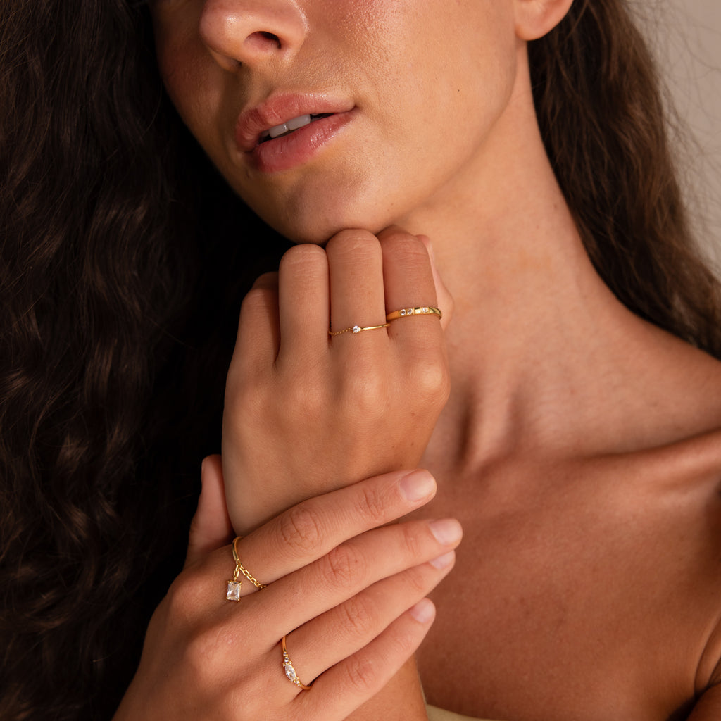 Woman with wavy hair showcases gold rings, including the Diamond Morse Code Ring in Rose Gold with a clear gemstone and hidden message, against a neutral background.