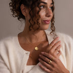 Woman with curly hair wearing gold jewelry, including the Baby Hand & Footprint Necklace, and a white cardigan, looking to the side with a gentle smile.