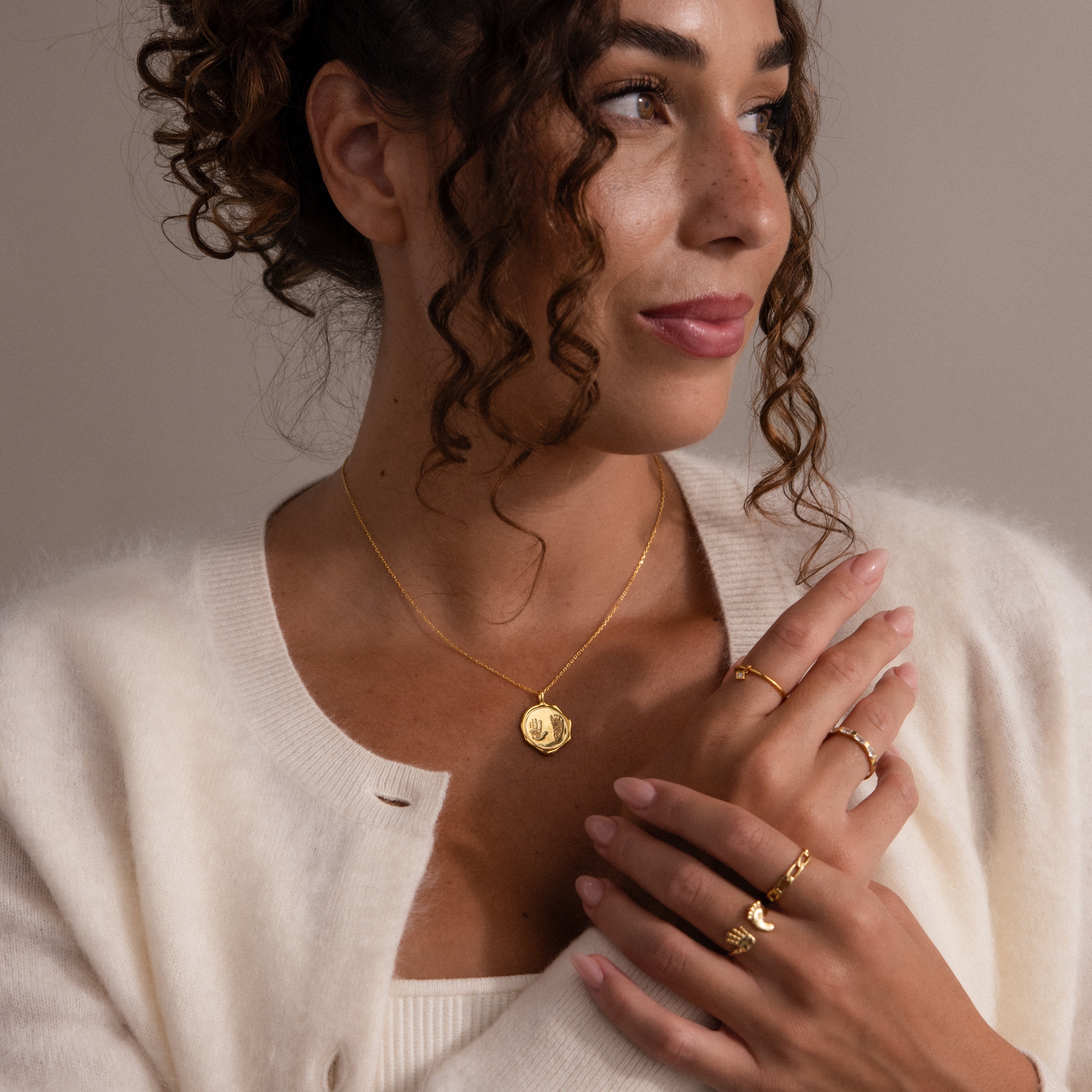 Woman with curly hair wearing gold jewelry, including the Baby Hand & Footprint Necklace, and a white cardigan, looking to the side with a gentle smile.