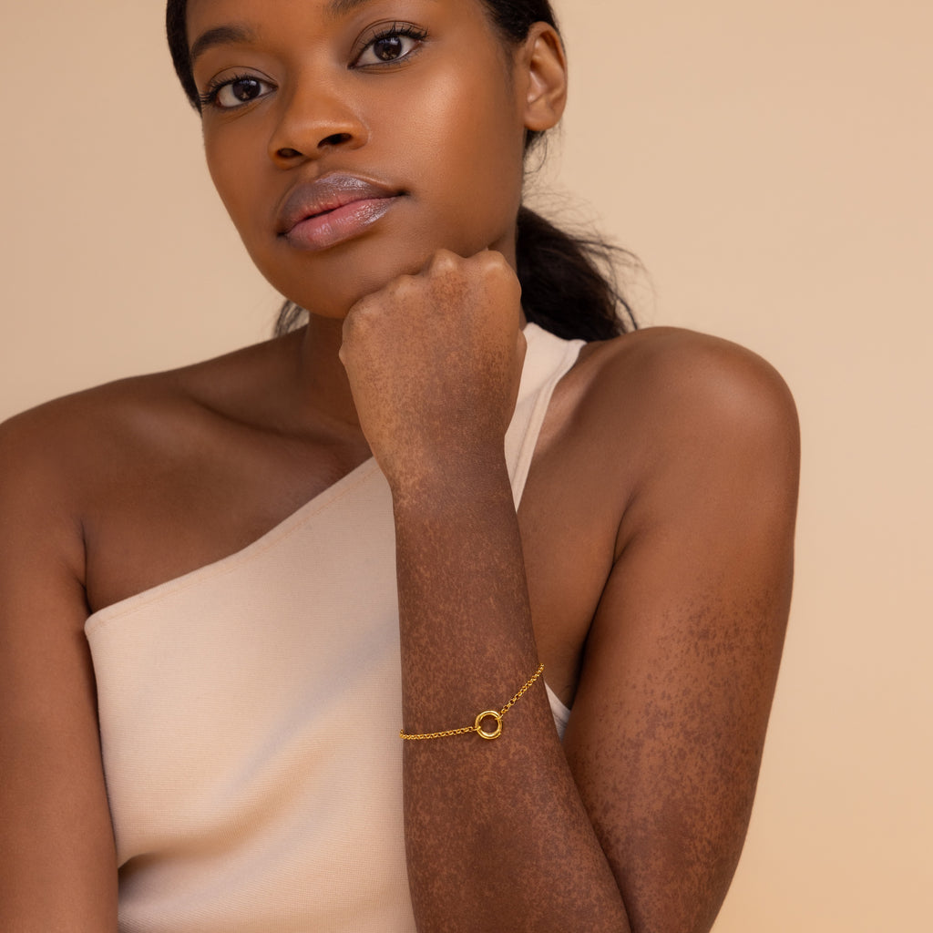 A woman in a one-shoulder top poses with her chin on her hand, highlighting the elegant Rolo Circle Charm Bracelet, featuring a detachable hinged circle for adding charms and personalizing your look.