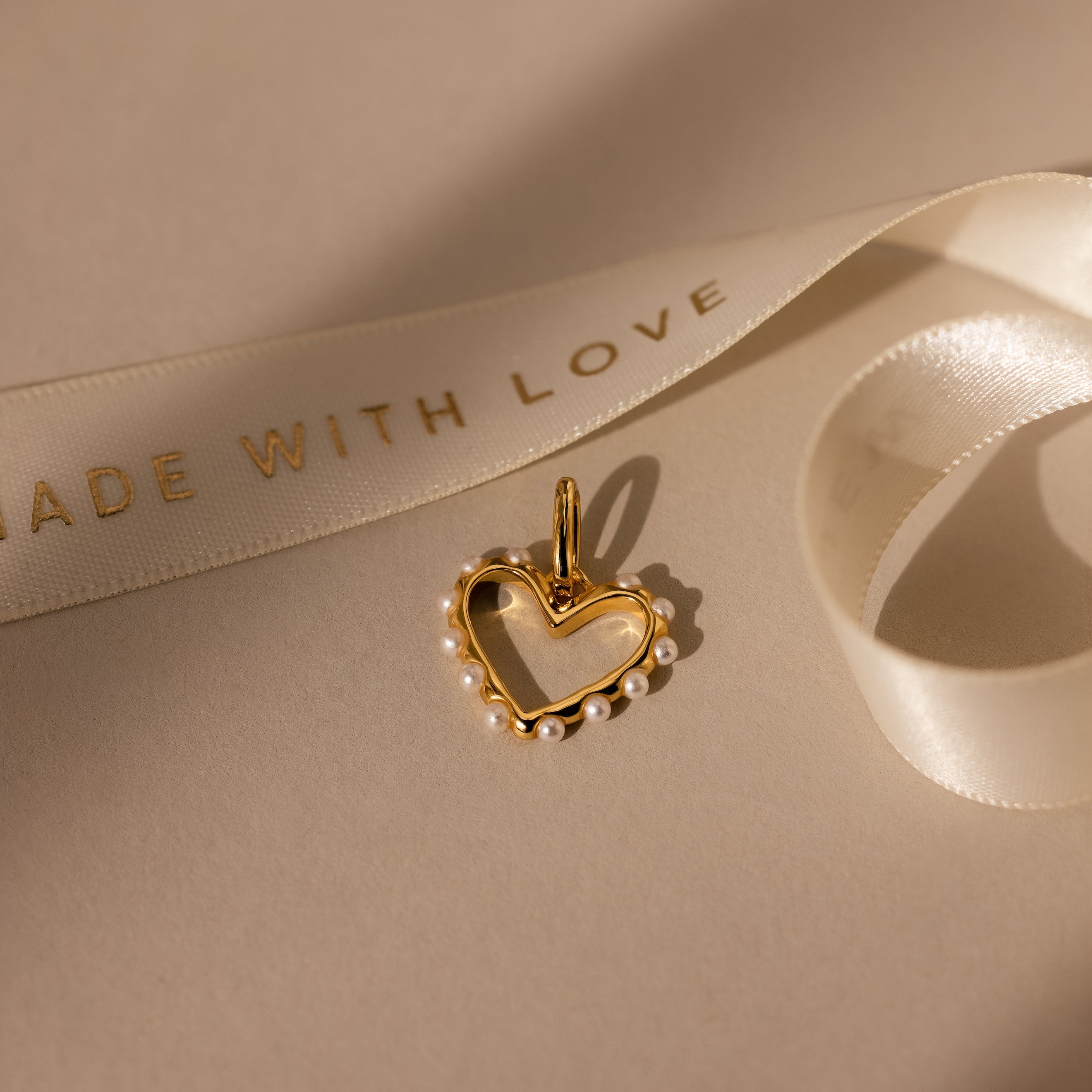 The Classic Pearl Heart Charm in 18K Gold, adorned with small pearls, is displayed beside a white ribbon printed with MADE WITH LOVE.