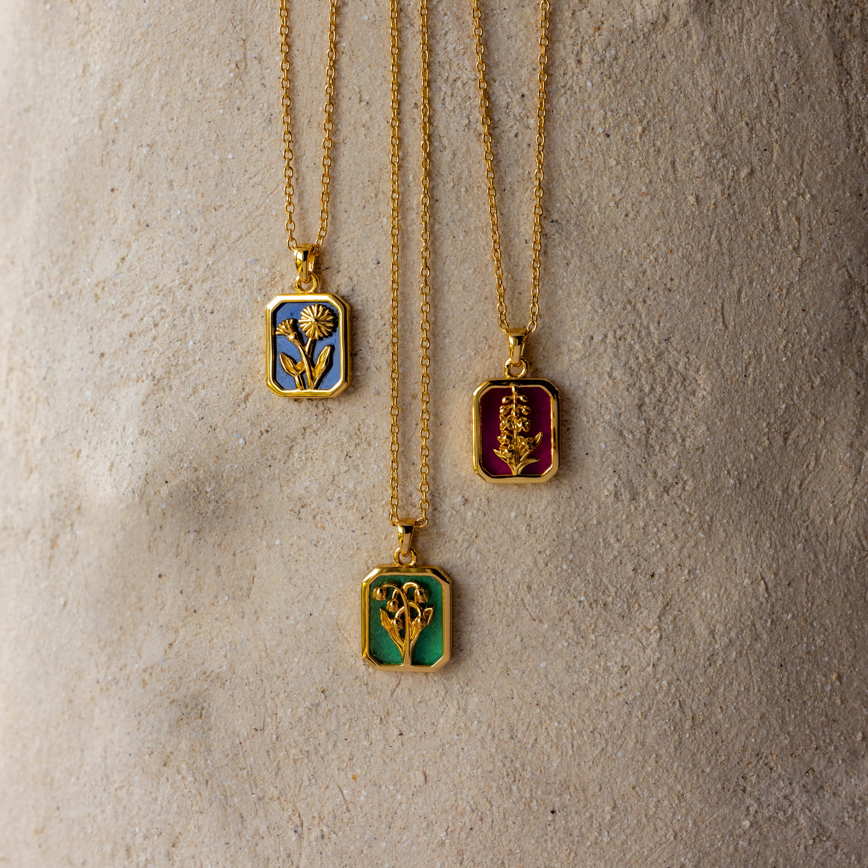 The Ruby Birth Flower Charm Necklace features a gold floral pendant with delicate ruby red enamel, resting elegantly on a stone surface and capturing timeless beauty.