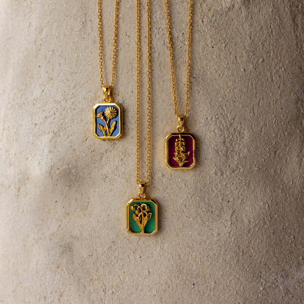The Sapphire Birth Flower Charm Necklace features a gold chain with a colorful rectangular pendant adorned with floral designs, elegantly displayed on a textured beige surface—an enchanting addition to any jewelry collection.