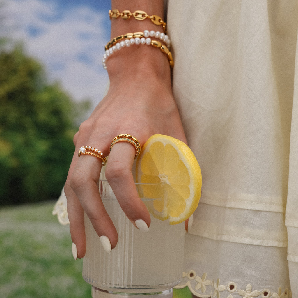 A hand wearing the Pearl Duo Ring and gold jewelry holds a glass with lemon, paired with a cream dress outdoors.