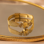 The Diamond Mesh Timepiece features a gold mesh band, round face, and diamond accents, shown on a reflective gold tray.