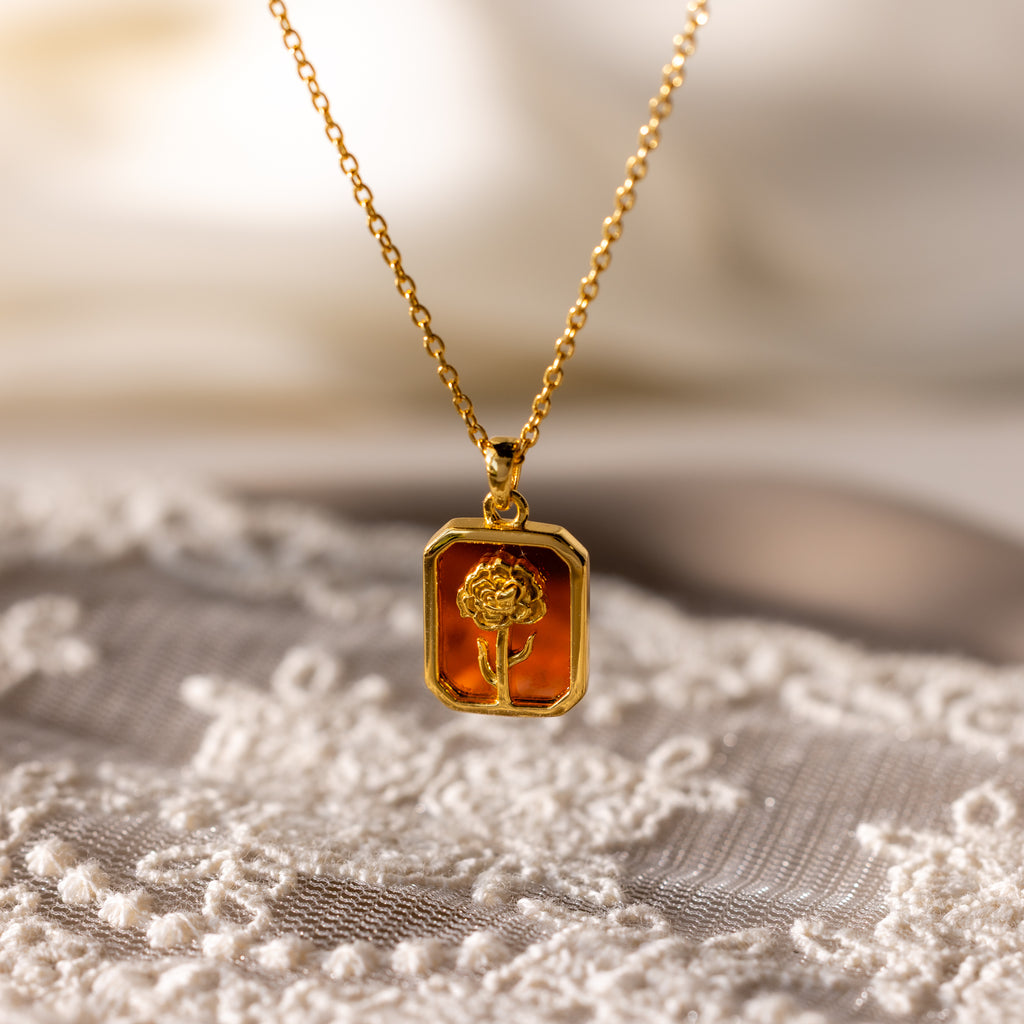 Garnet Birth Flower Charm Necklace with a rectangular pendant featuring a raised flower design, displayed on lace fabric.