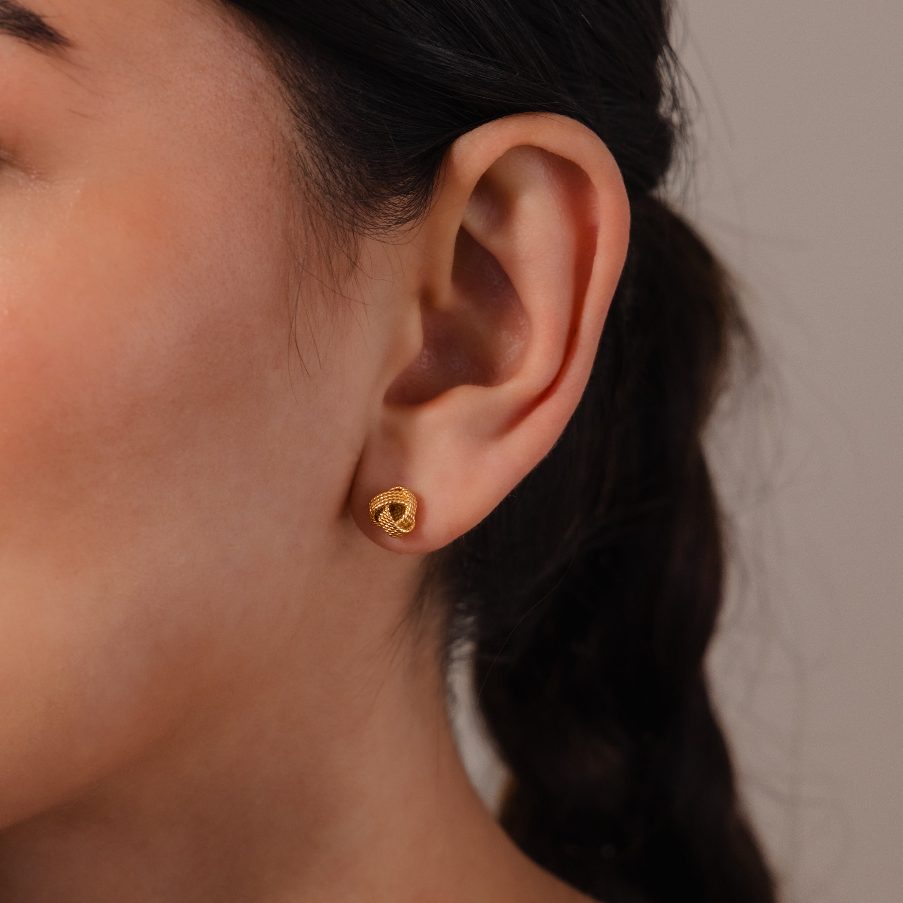 Close-up of a woman's ear wearing Rope Knot Studs, small round gold earrings; her hair is styled in a braid for an elegant, classic look.