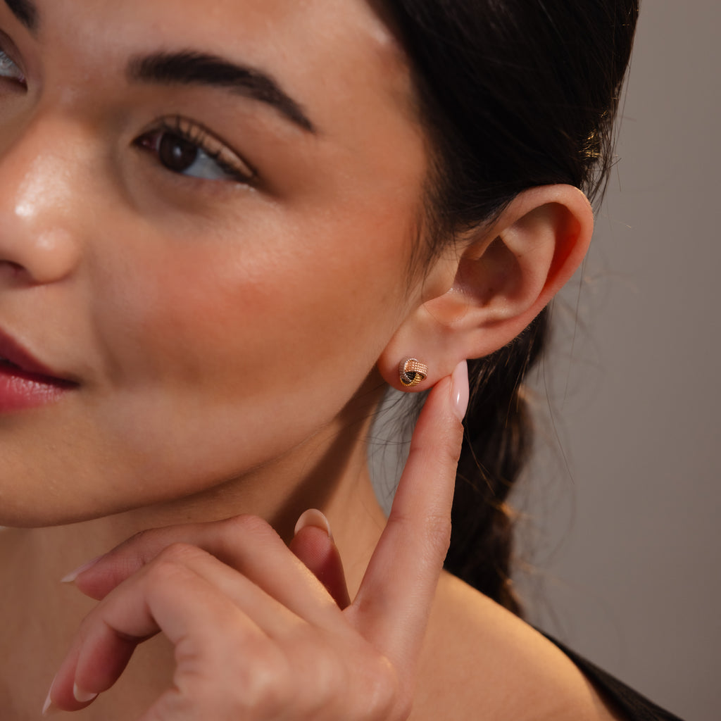 A woman with braided hair gently points to her Rope Knot Studs in Mixed Metal, her soft smile drawing attention to the elegant earrings.