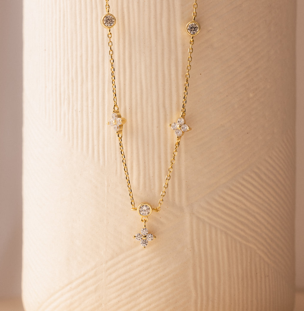 The Ivy Diamond Station Necklace in 18K Gold features small round and star-shaped diamond accents, displayed against a textured beige background.