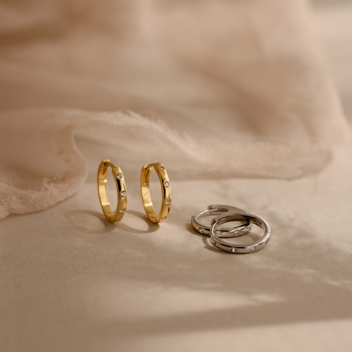Three minimalist metal huggies with tiny clear stone accents, including two gold rings and one silver ring, arranged on a soft beige fabric background.
