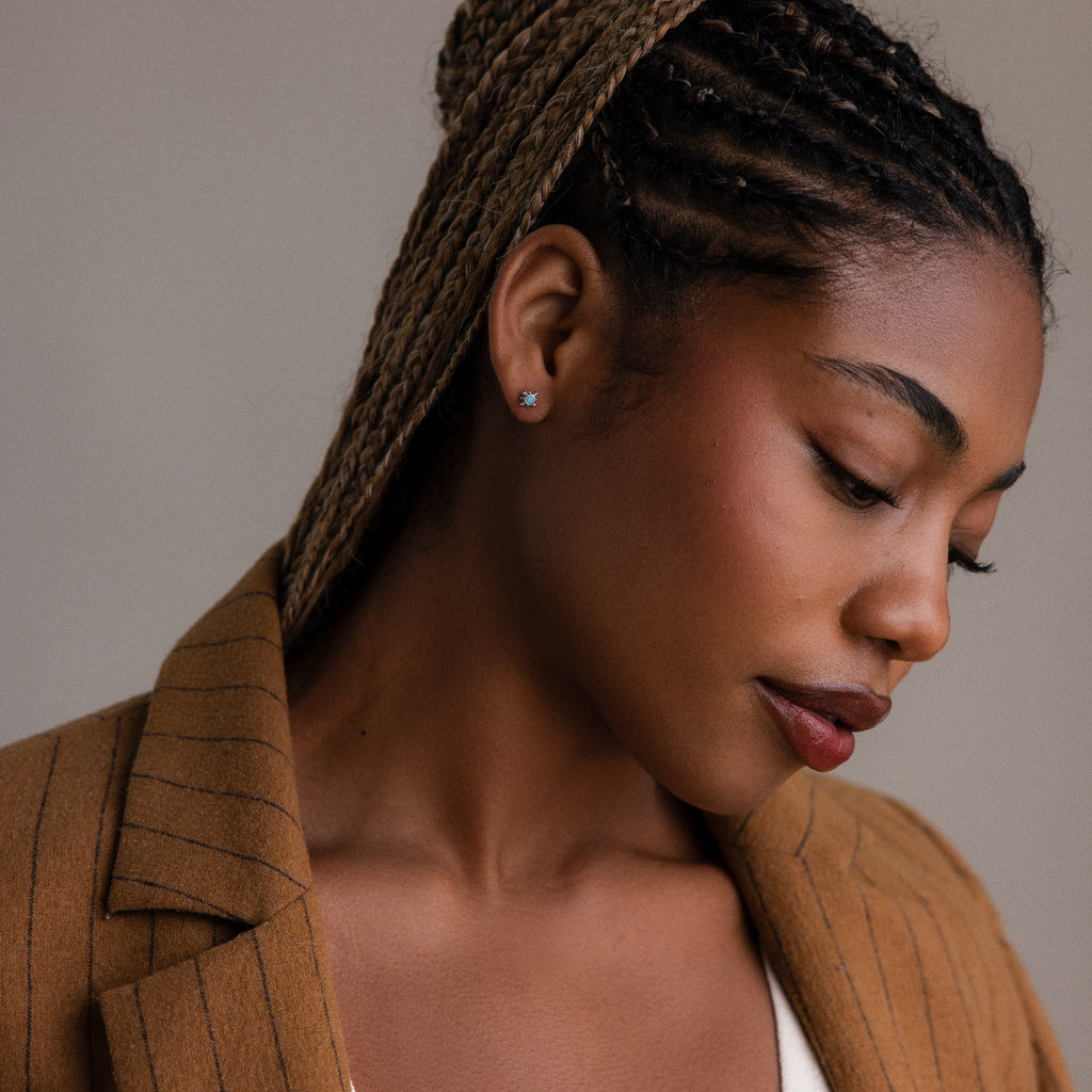 Model wearing dainty turquoise gemstone stud earrings in a silver setting, styled as minimalist everyday jewelry with a warm neutral outfit.