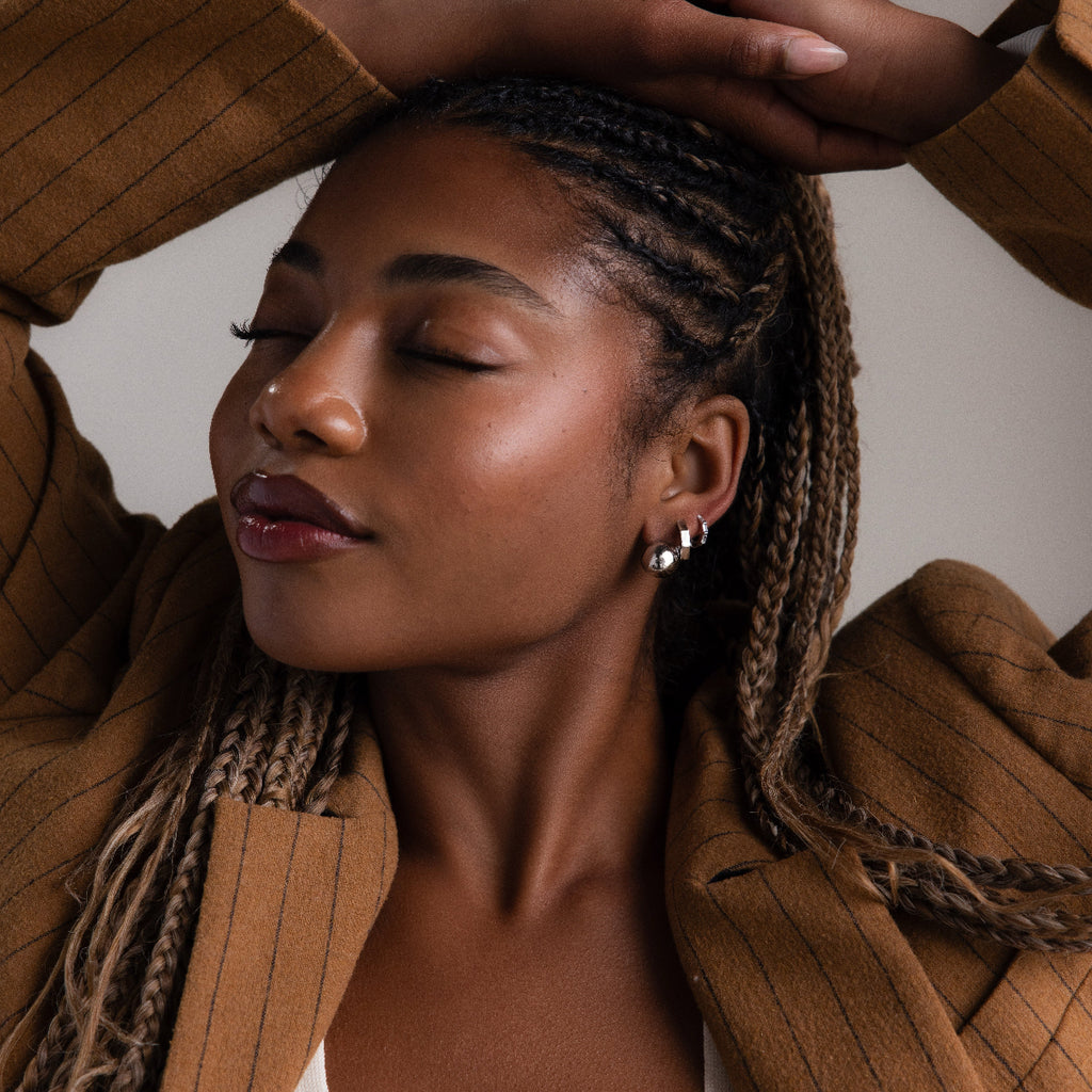 Woman with braided hair wearing a brown coat and multiple silver earrings against a neutral background