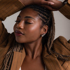 Woman with braided hair wearing a brown coat and multiple silver earrings against a neutral background