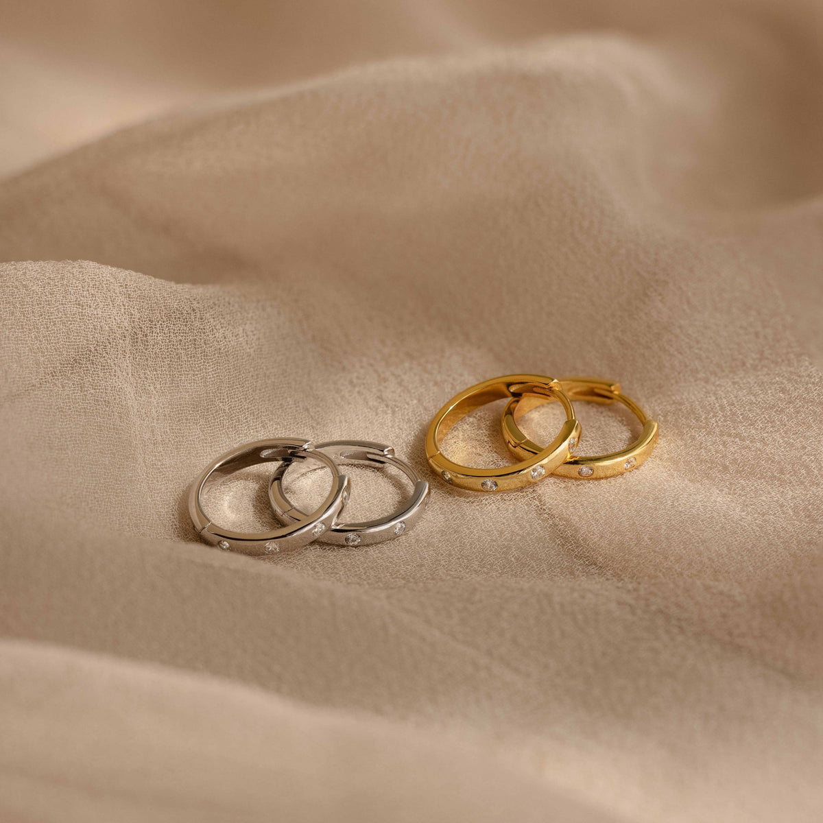 Classic Diamond Sprinkle Huggies shown in silver and gold finishes resting on a soft beige fabric background.