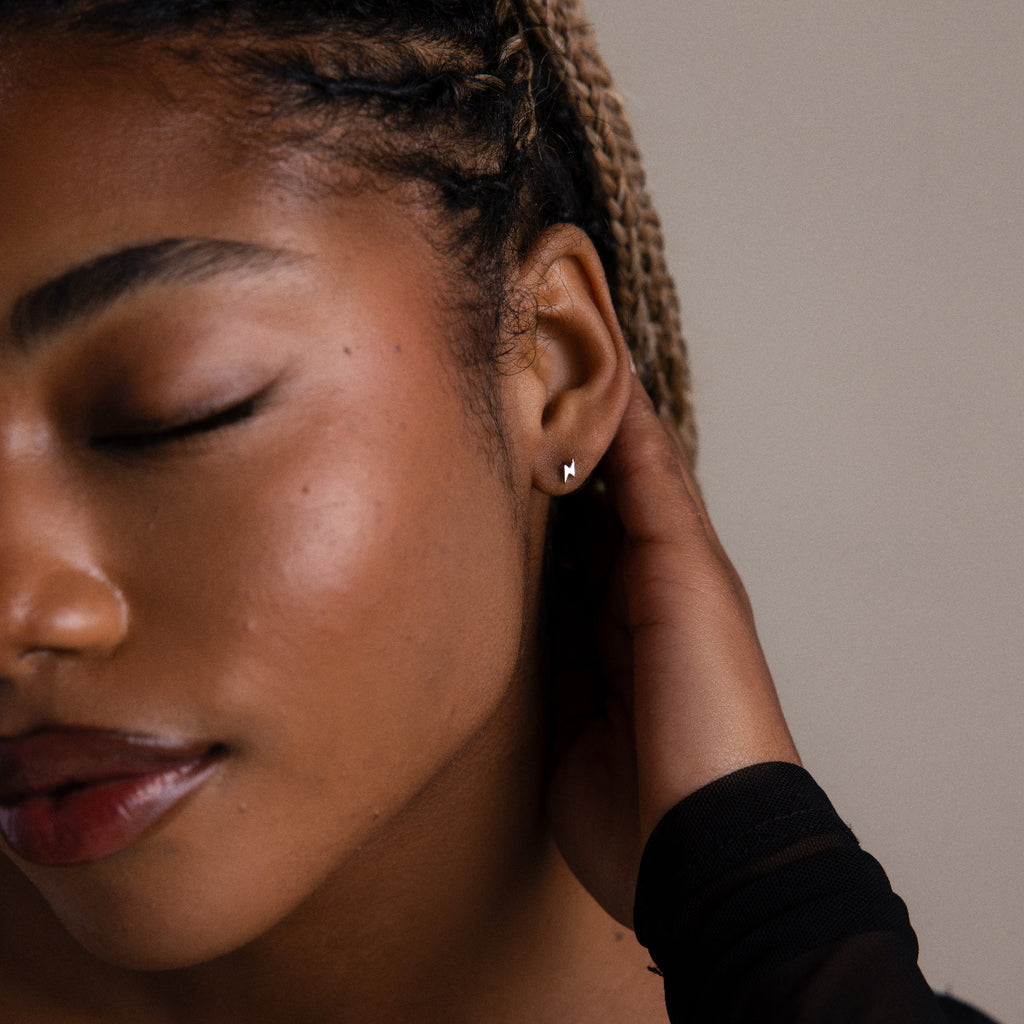 Close-up of a woman with braided hair touching her ear, wearing a small silver lightning bold stud earring.