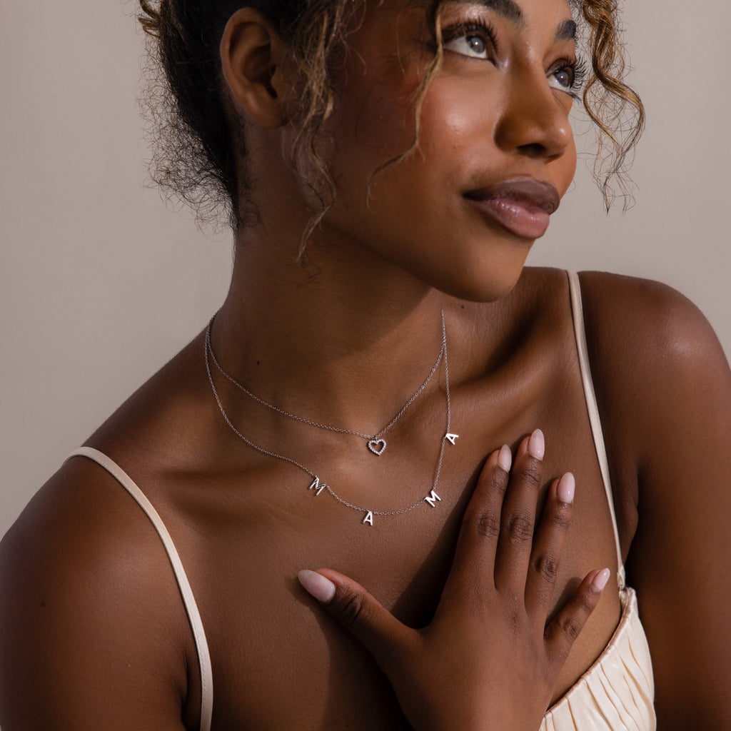 Woman in a beige dress touches her chest, wearing layered “MAMA” necklaces accented by the Forever Pave Heart Necklace and a delicate heart charm.