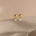 The Heart Pearl Drop Earrings in 18K Gold feature gold heart-shaped studs with small white pearl dangles, elegantly displayed on a neutral textured surface.