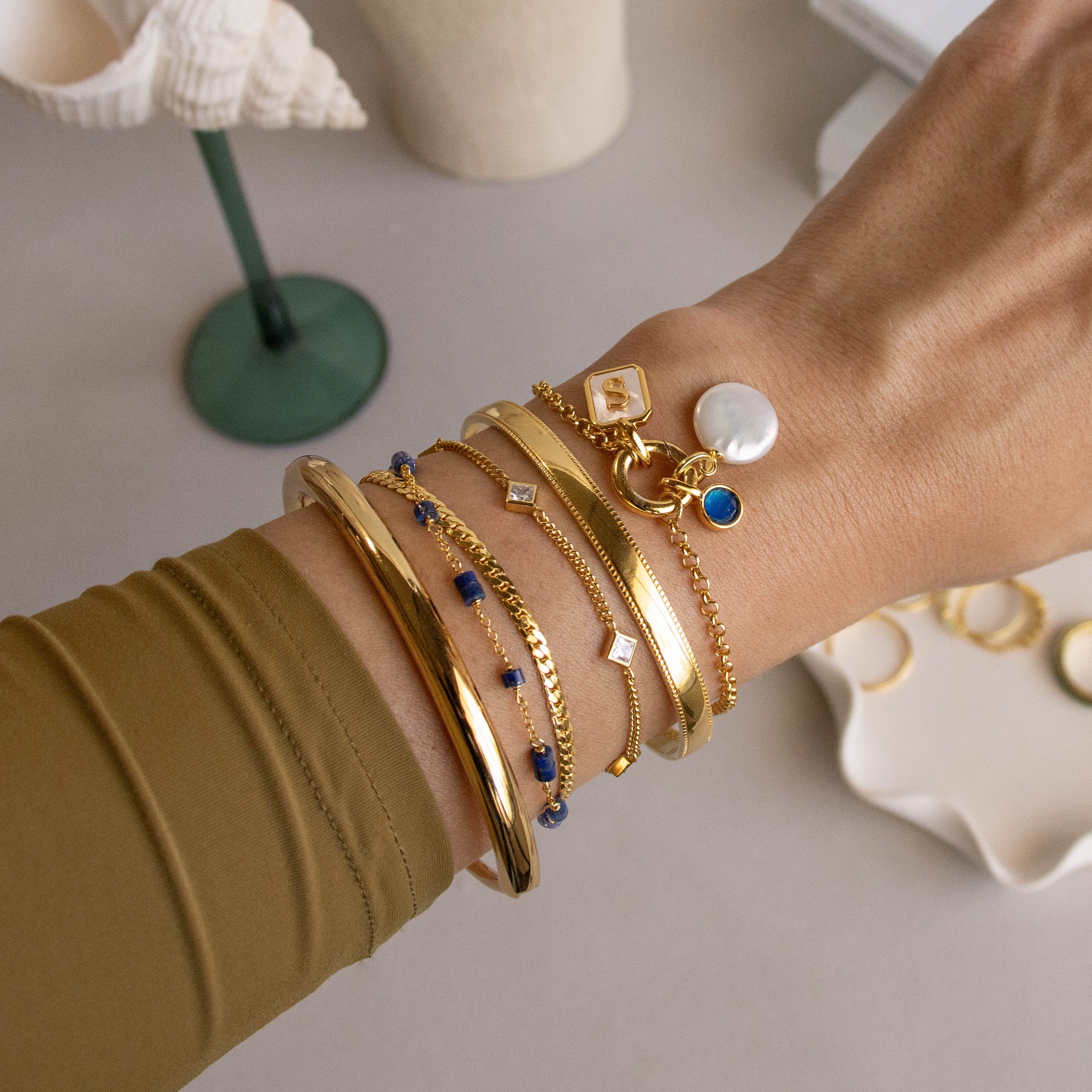A hand wearing layered gold bangles, the Alyssa Cuff Bracelet, and bracelets with blue, white, and pearl charms.