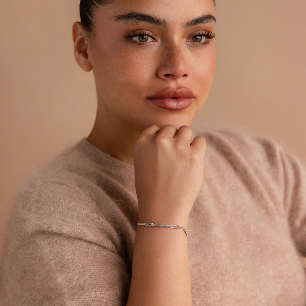 A woman in a beige sweater rests her chin on her hand, wearing a delicate Love Knot Bracelet in White Gold—a thoughtful gift for couples—set against a neutral background.