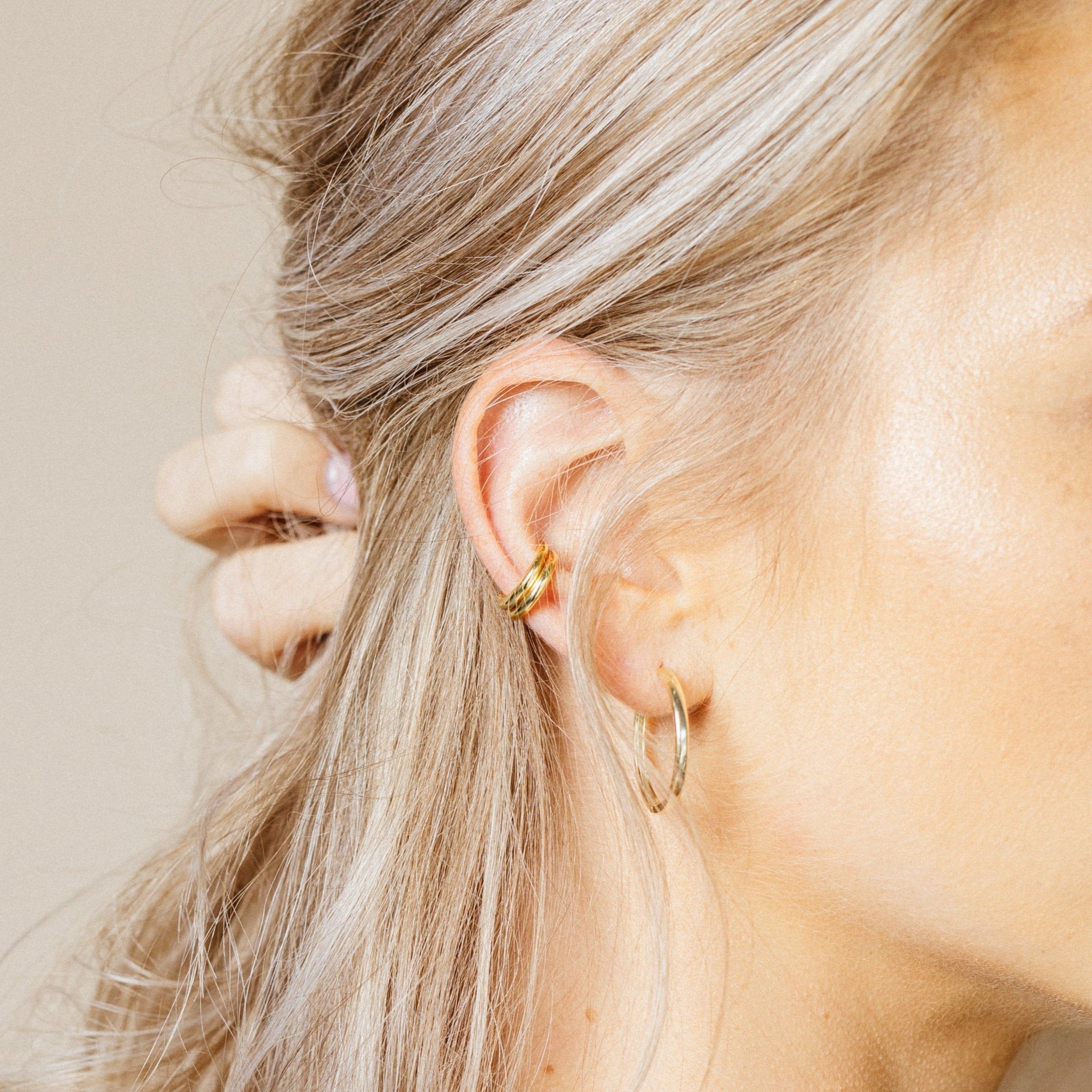 Close-up of a person wearing 2 stacked gold cuff earrings with a gold hoop earring in a neutral background
