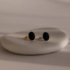 Two Midnight Hour Onyx Rings—one gold, one silver, each with an adjustable band—are displayed on a smooth stone surface for a vintage-glam touch.