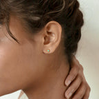 Close up of a model wearing a dainty gold stud earring with a small turquoise gemstone, styled as minimalist everyday jewelry with a soft natural look.