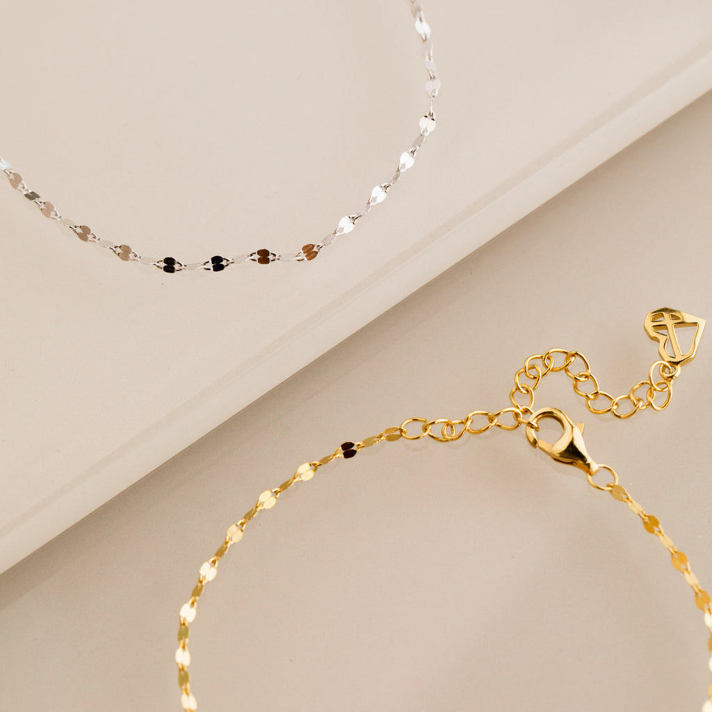 The Josephine Chain Bracelet, shown in silver and gold on a beige surface, features a delicate anchor link design.