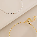 The Josephine Chain Bracelet, shown in silver and gold on a beige surface, features a delicate anchor link design.