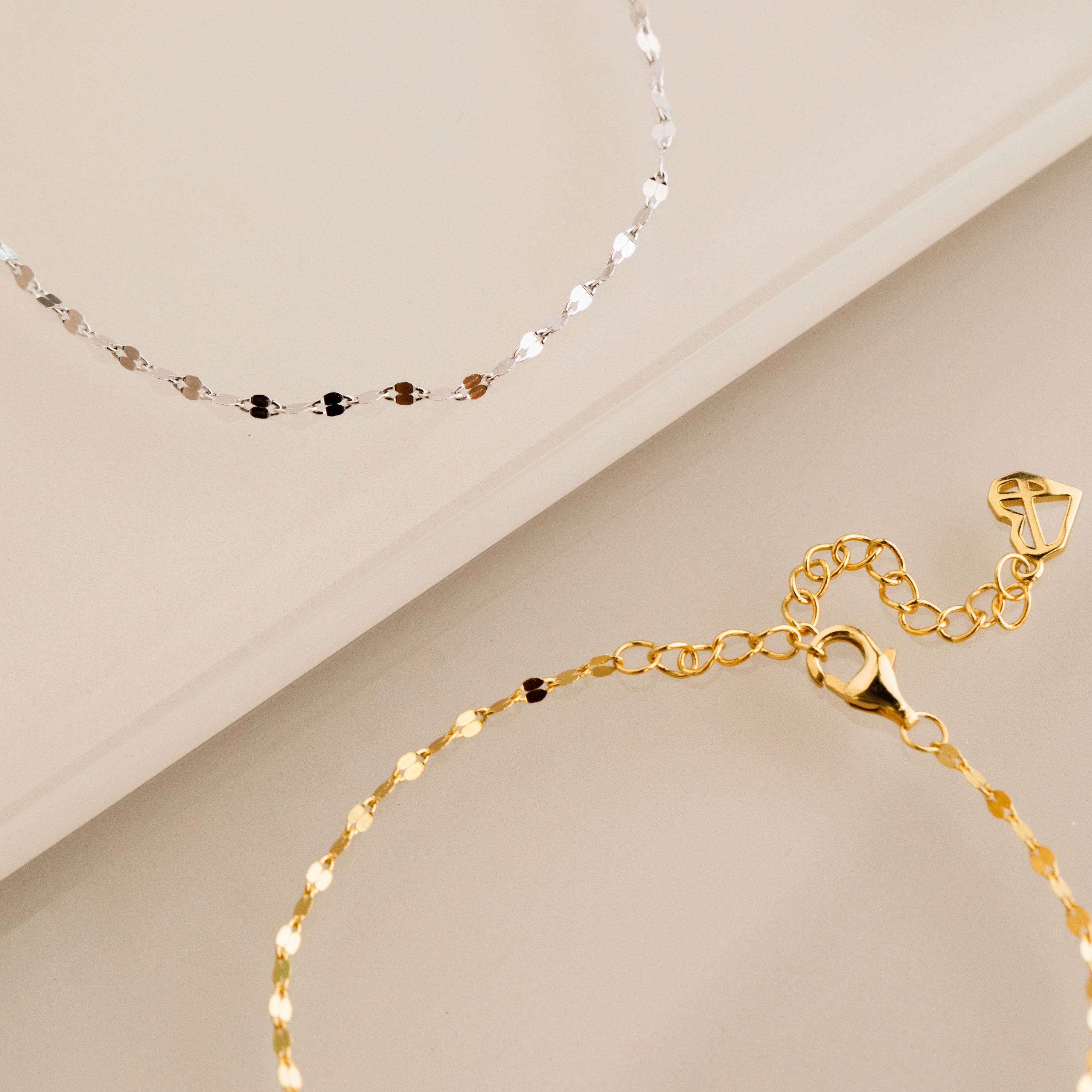 The Josephine Chain Bracelet, shown in silver and gold on a beige surface, features a delicate anchor link design.
