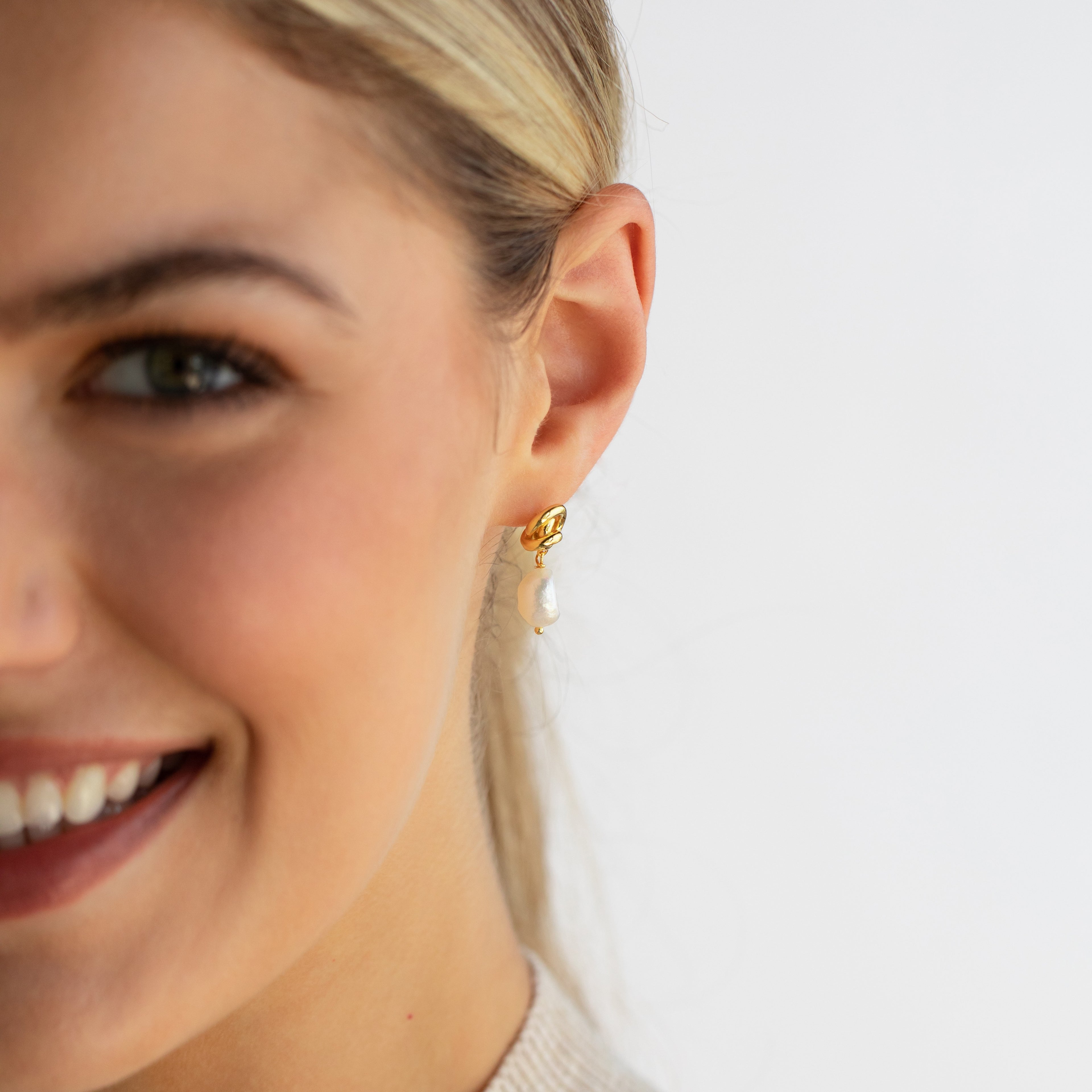 A smiling woman wears Knot Pearl Drop Earrings in 18K Gold—a delicate gold and pearl drop design with a subtle Celtic knot—shown from cheek to ear against a white background.