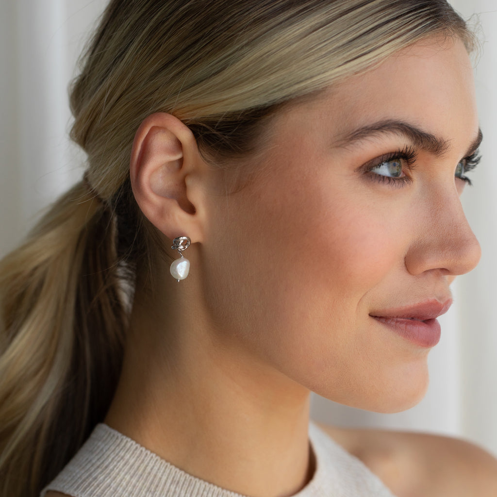 A blonde woman with her hair in a ponytail smiles softly, looking to the right while wearing the Knot Pearl Drop Earrings.