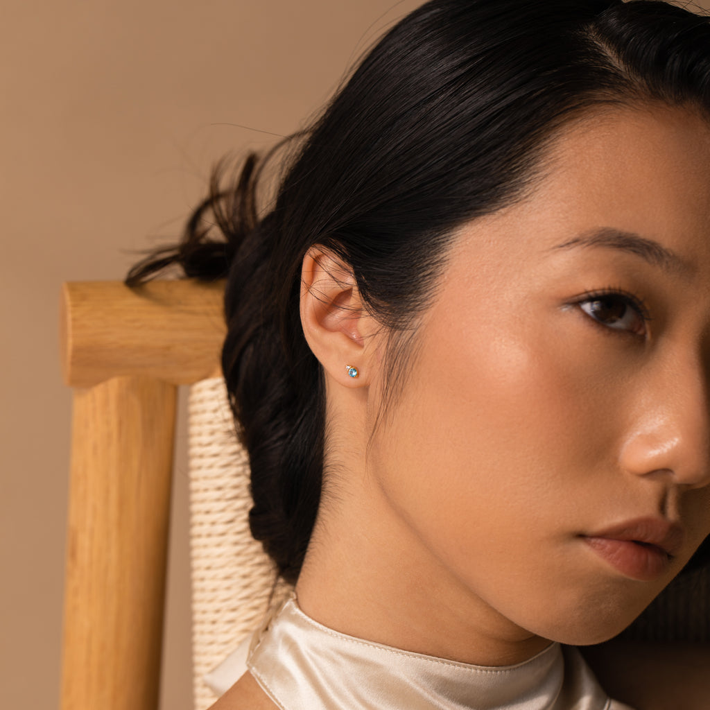 Close-up of a woman with smooth skin, wearing a white satin top and Raindrop Emerald Studs, sitting on a wooden chair.