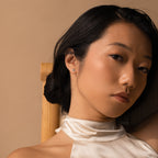A woman with smooth skin and dark hair sits on a wooden chair, wearing a white satin halter top and the Raindrop Pearl Studs.