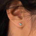 Close-up of an ear adorned with Raindrop Topaz Studs: a small blue topaz earring paired with a tiny clear diamond stud above it.