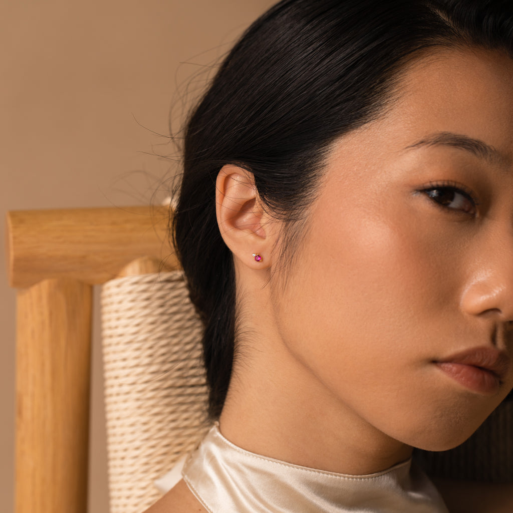 Woman with smooth skin wears Raindrop Ruby Studs, seated on a wooden chair against a beige background.