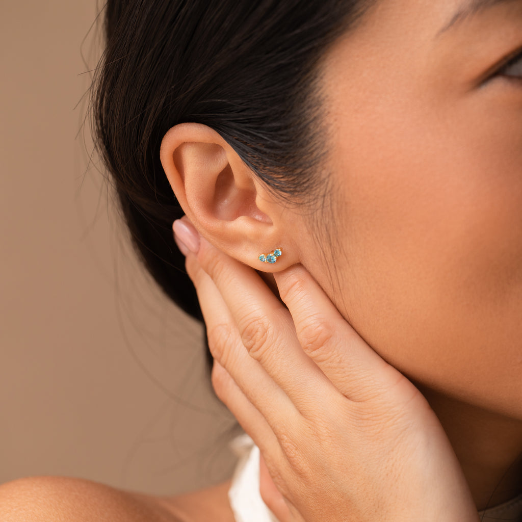 A woman touches her ear, showcasing the Bella Birthstone Studs—a delicate turquoise earring thats a thoughtful gift for mothers and daughters.