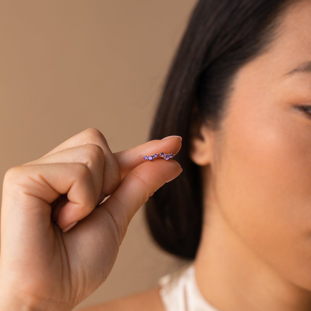 A woman holds the Bella Amethyst Studs—small, curved earrings with purple gems—close to her face, making them a perfect personalized gift for mothers and daughters.