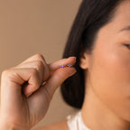 A woman holds the Bella Amethyst Studs—small, curved earrings with purple gems—close to her face, making them a perfect personalized gift for mothers and daughters.
