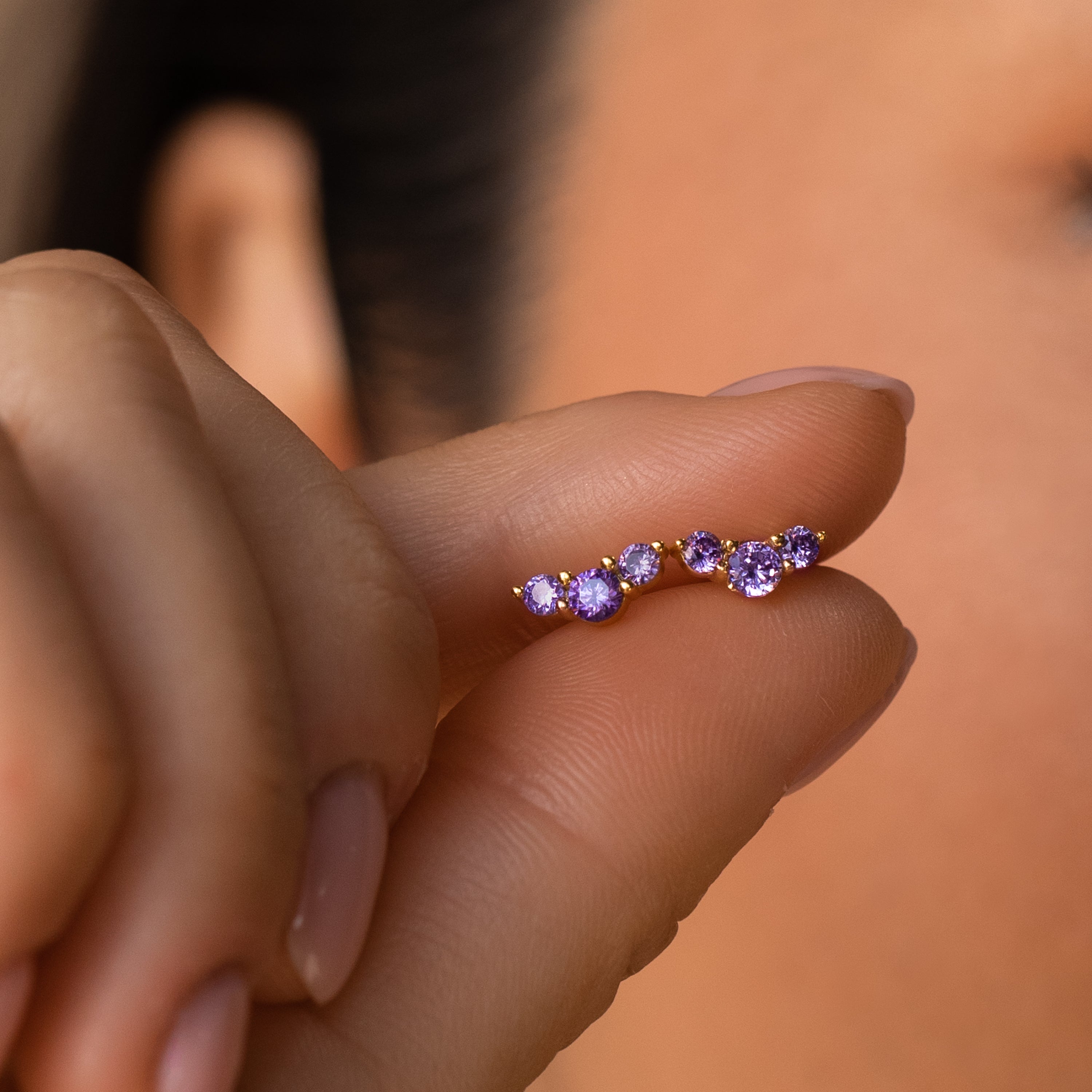 A hand holds a Bella Birthstone Stud earring with five small purple gemstones near a face against a neutral background—ideal as personalized jewelry or a meaningful gift for mothers and daughters.
