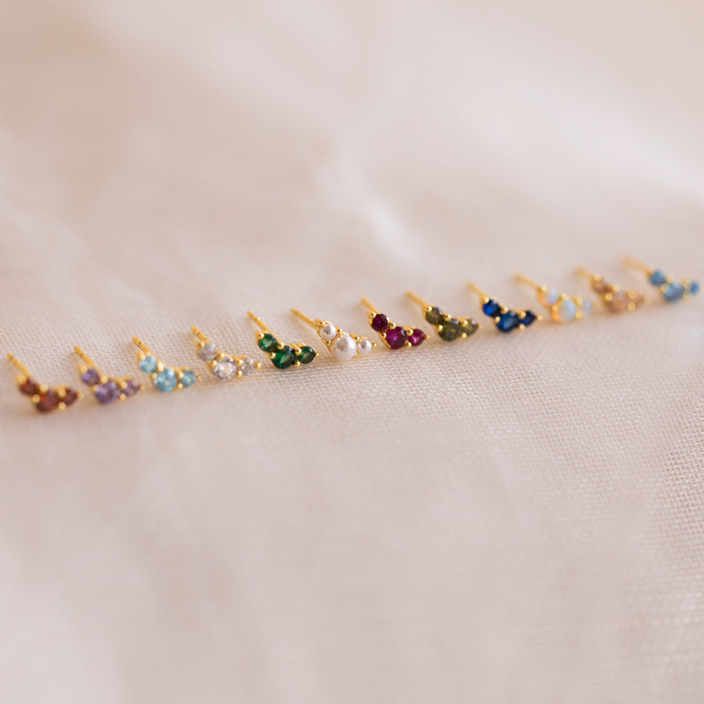 A row of Bella Aquamarine Studs with gold settings is arranged on a soft, light fabric background—perfect as personalized jewelry or a thoughtful gift for mothers and daughters.