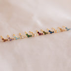 Bella Emerald Studs—small, vibrant green birthstone studs set in gold—are displayed on white fabric, making an ideal personalized jewelry gift for mothers and daughters.