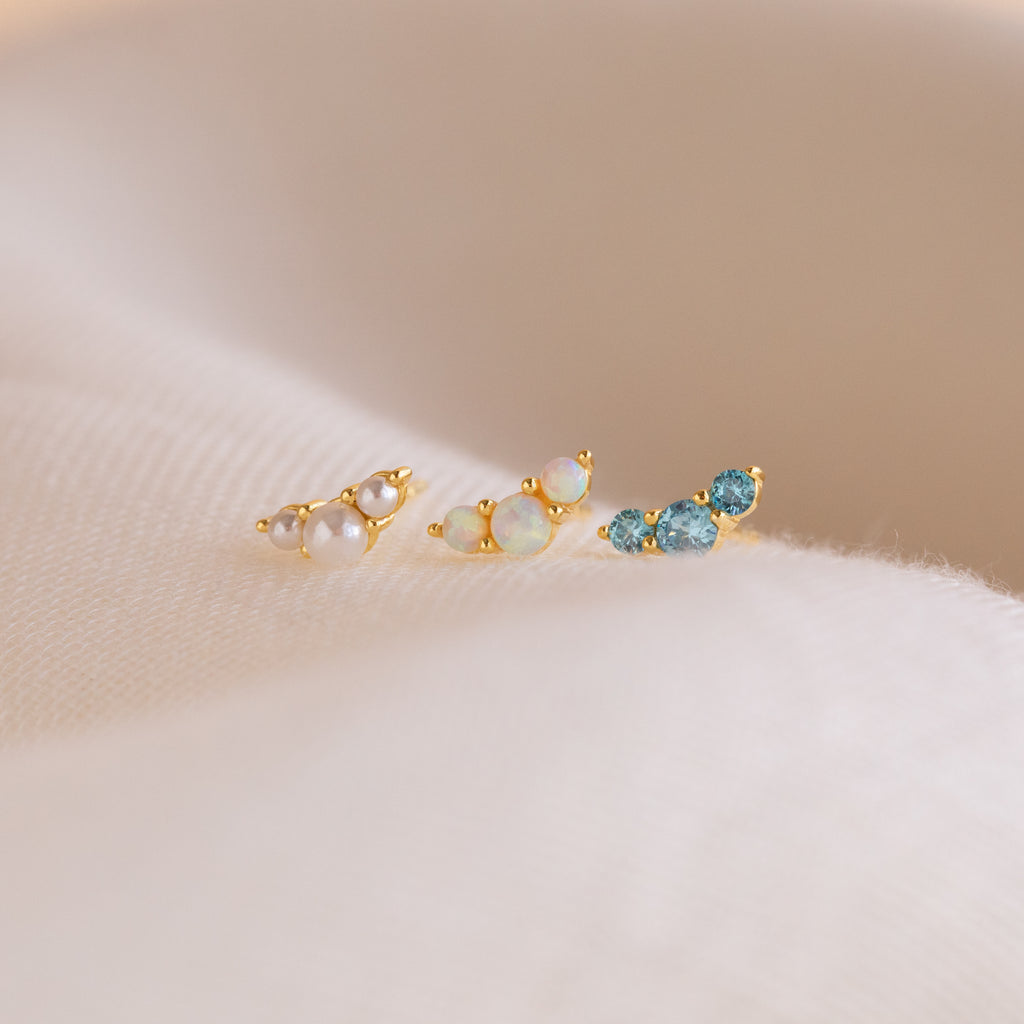 The Bella Birthstone Studs, a set of three gold stud earrings with pearls, opals, and blue gems on soft beige fabric, make a perfect personalized gift for mothers and daughters.
